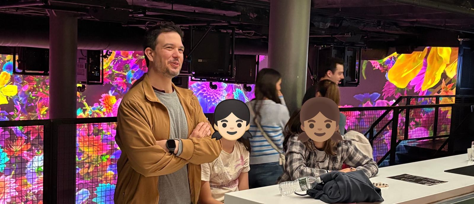 Daniel standing at the XR Bar with his daughter and her friend, AI-generated floral imagery and the main PIXELBLOOM show visible on screens behind them
